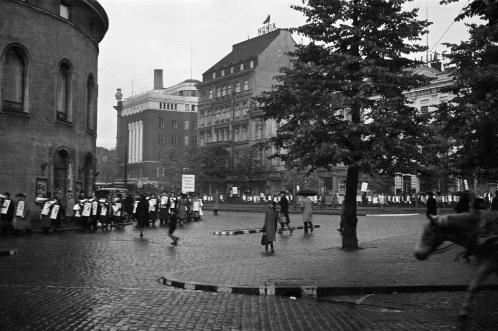 Jehovan todistaja, insinööri Kaarlo Hartevan esitelmää mainostava kulkue. Miehiä ja naisia mainos- ja tekstikylttien kanssa kääntymässä Mannerheimintieltä Pohjoiselle Esplanaadikadulle (=Pohjoisesplanadi). Vasemmalla Ruotsalainen teatteri, taustalla Erottajankatu 4, Mannerheimintie 2, 4.