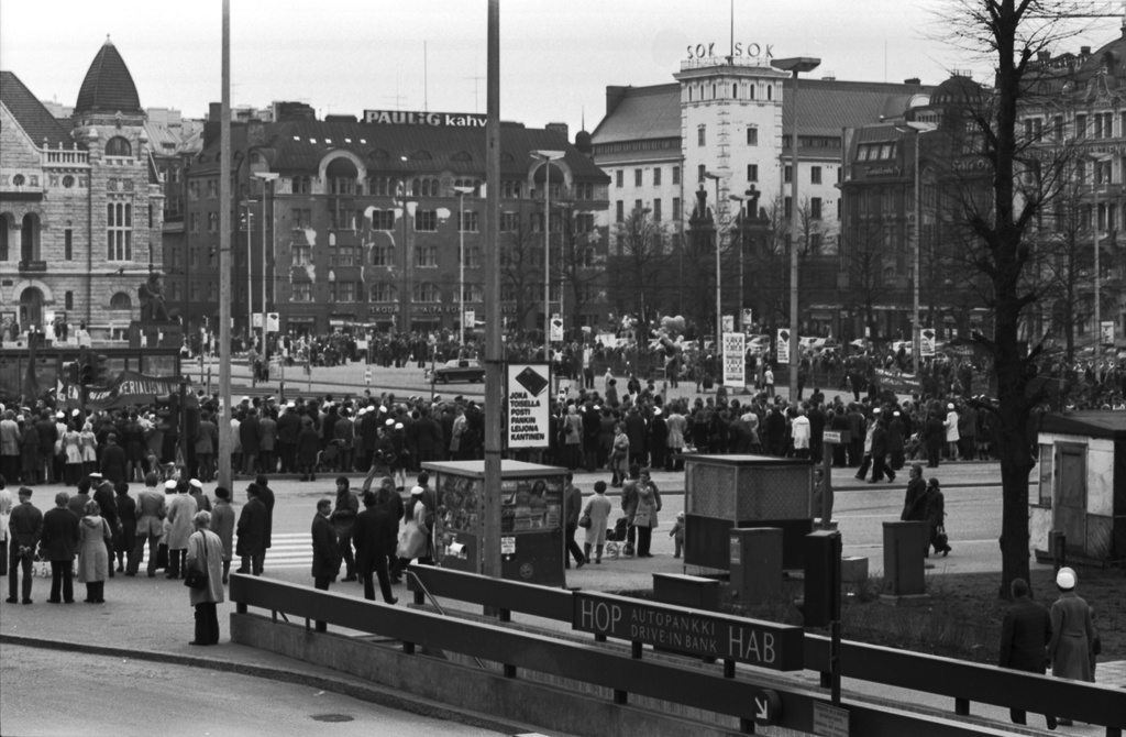 Kaivokatu. Runsaslukuinen joukko ihmisiä katujen laidalla seuraamassa työväen vappukulkuetta, joka on kiertymässä Rautatientorin ympäri Mikonkadulta Kaivokadulle 1.5.1972.  Etualalla portaat asematunneliin ja sisäänajo Helsingin Osakepankin drive-in autopankkiin Keskuskatu 7. Kaivokadun jalkakäytävällä lehti- makkara- ja kukkakioskit. Näkymä Makkaratalolta Vilhonkadun suuntaan.