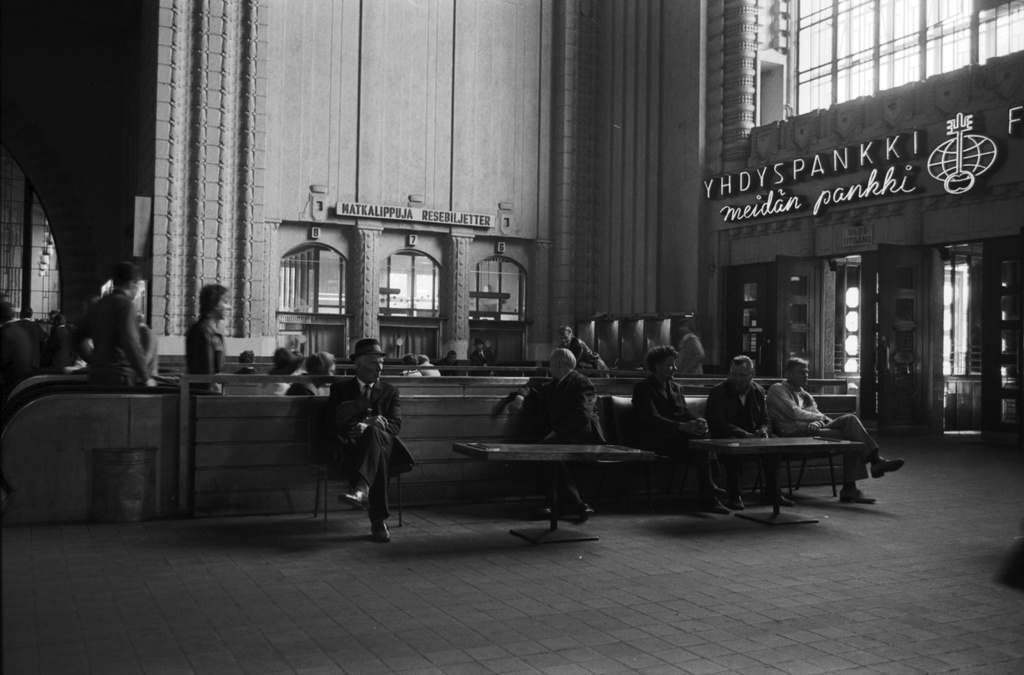 Helsingin rautatieasema. Ihmisiä Helsingin rautatieaseman Kaivokadun puoleisessa asemahallissa. Ulko-ovien päällä Yhdyspankki meidän pankki -neonvalomainos.