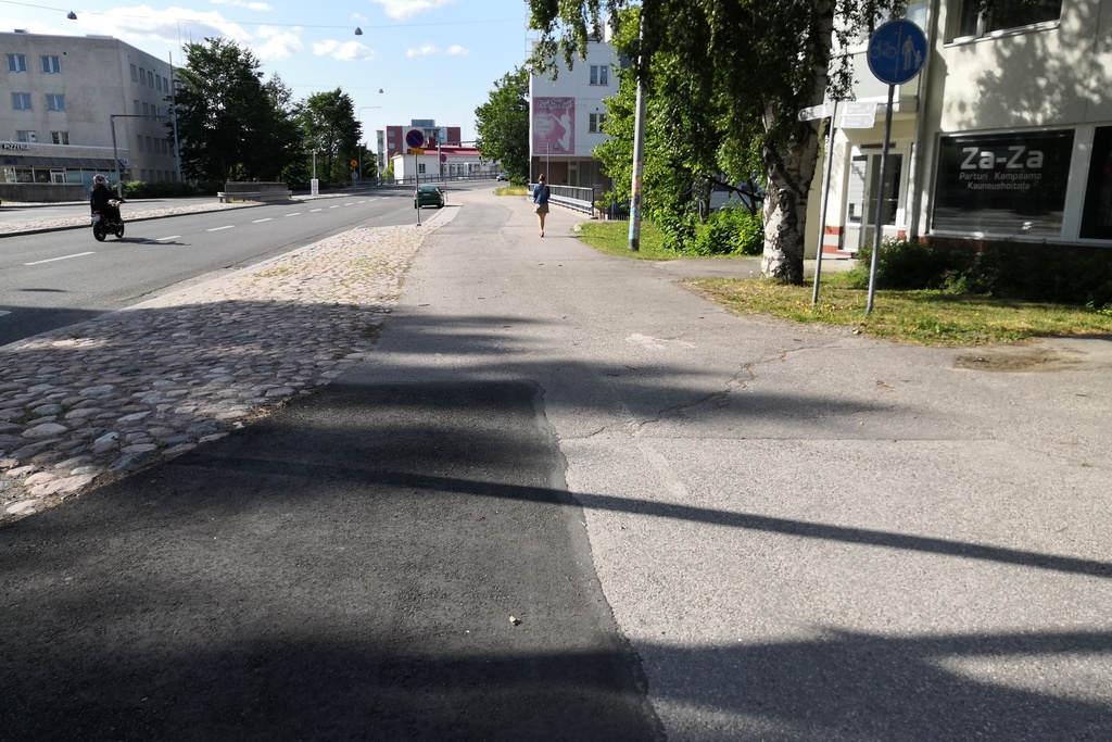 Malmi Ylä-Malmi. Kolme naista, todennäköisesti postinjakajia, polkupyörineen Kirkonkyläntiellä. Syksyinen näkymä auton ikkunasta. Oikella talo, jossa ravintola Malmi Kesti ja R-kioski. Kioskin luukulla mies asiomassa. rephoto