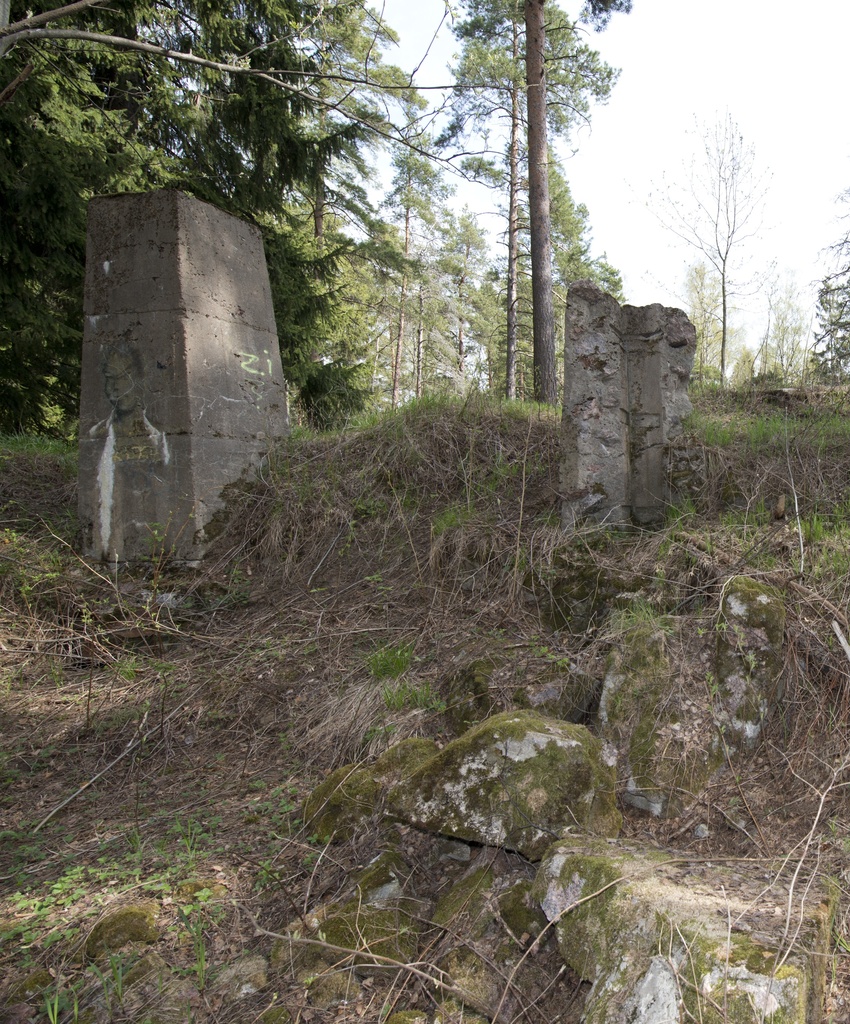 Myllypuro. Ensimmäisen maailmansodan aikainen maalinnoitus, tykkipatteri 65 Myllypurossa. Kuvassa näkyy kaksi pohjoisimman suojahuoneen viidestä betonisesta savupiipusta tai hormista. Vasemmanpuolimmainen hormi on ehyt ja sen kylkeen on maalattu ihmishahmoa esittävä vihertävä graffiti. Oikeanpuolimainen hormi on pahasti vaurioitunut, vain yksi hormin kulmista on enää pystyssä. Kulmissa näkyy painanteina hormia tukeneista vahvikkeista. Kuvaussuuntana pohjoinen.