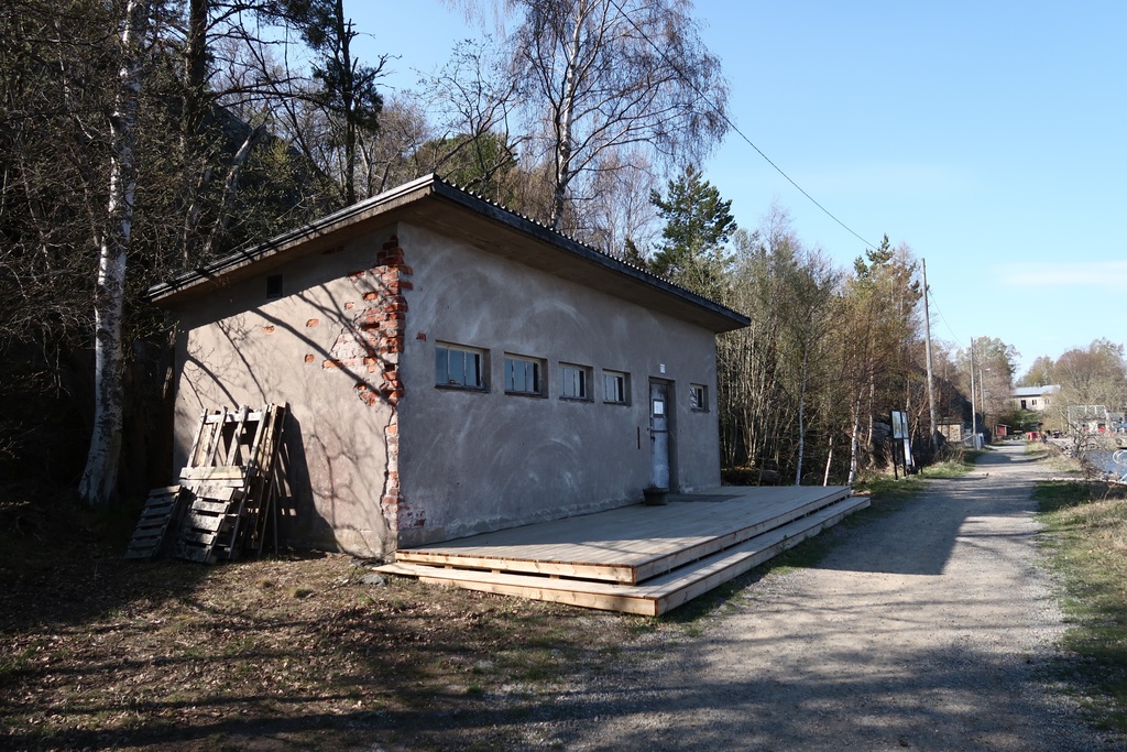 Näkymä Vallisaaren itärantaa kulkevalle tielle. Se on retkisataman tuntumassa jo suurin piirtein Kuninkaansaarta vastapäätä. Punatiilinen rakennus vasemmalla liittyy saaren torpedoluolaston toimintaan. Rakennus on rapattu harmaalla laastilla, josta osa on karissut pois. Rakennuksen päätyä vasten nojaa lavoja ja julkisivun edustalle on rakennettu alusta. Äärimäisenä oikealla pilkottaa meri.