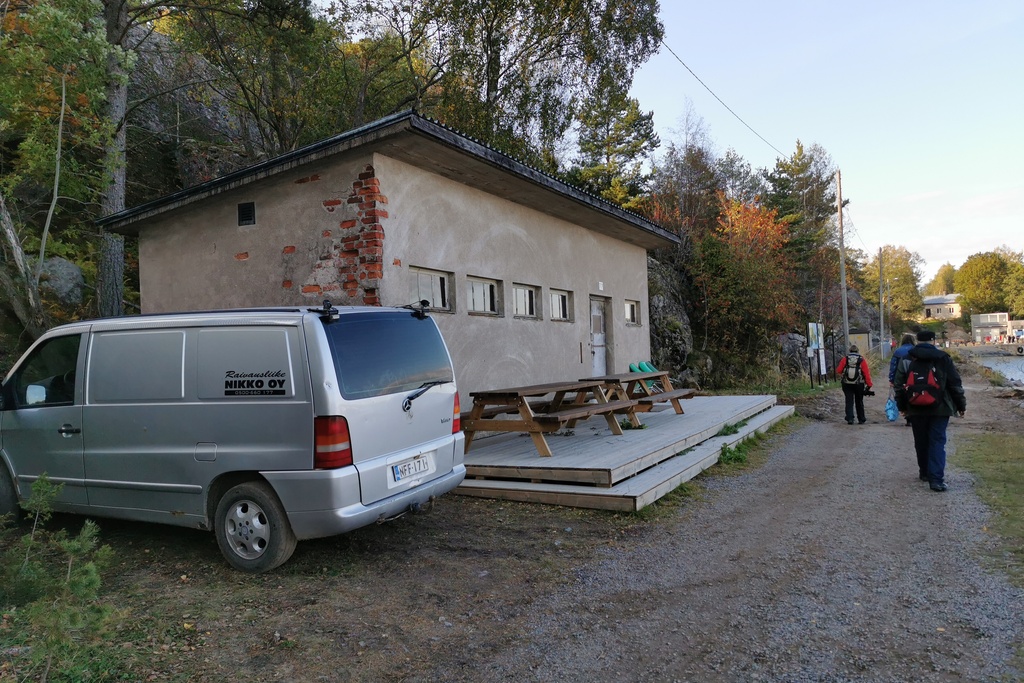 Näkymä Vallisaaren itärantaa kulkevalle tielle. Se on retkisataman tuntumassa jo suurin piirtein Kuninkaansaarta vastapäätä. Punatiilinen rakennus vasemmalla liittyy saaren torpedoluolaston toimintaan. Rakennus on rapattu harmaalla laastilla, josta osa on karissut pois. Rakennuksen päätyä vasten nojaa lavoja ja julkisivun edustalle on rakennettu alusta. Äärimäisenä oikealla pilkottaa meri. rephoto