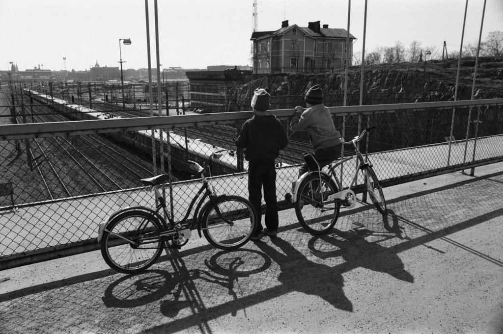Kaksi poikaa (Tuomas Heikkilä ja Aapo Rista) polkupyörineen katselemassa junien kulkua Linnunlaulun sillalla. Näkymä sillalta etelälounaaseen. Kalliolla Linnunlauluntie 12 puutalo.