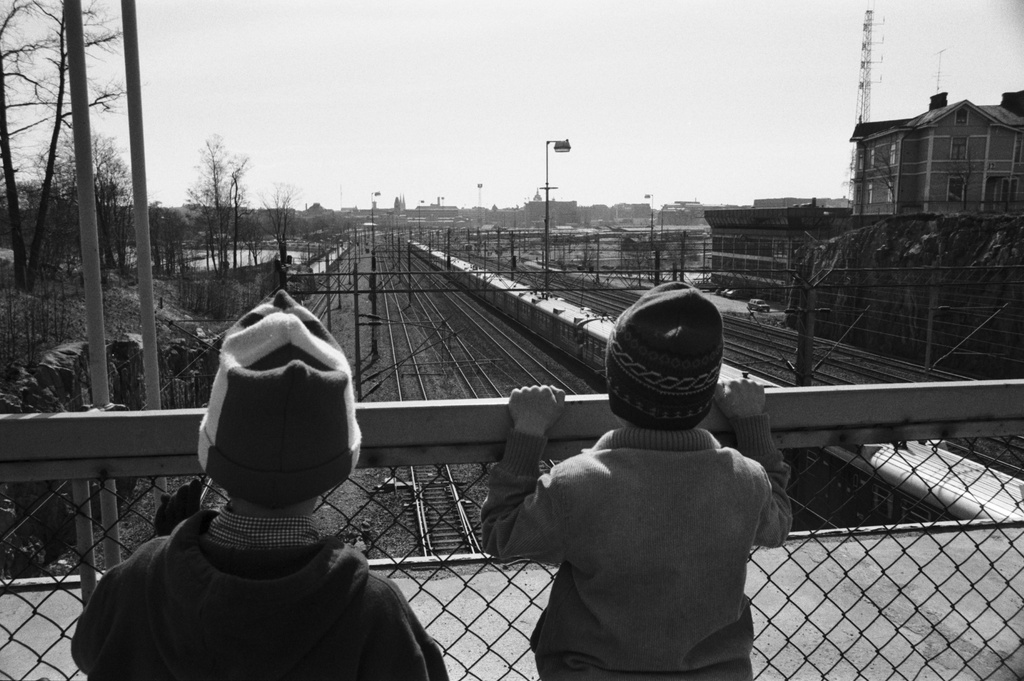 Kaksi poikaa (Tuomas Heikkilä ja Aapo Rista) polkupyörineen katselemassa junien kulkua Linnunlaulun sillalla. Näkymä sillalta etelään Helsingin rautatieasian suuntaan.