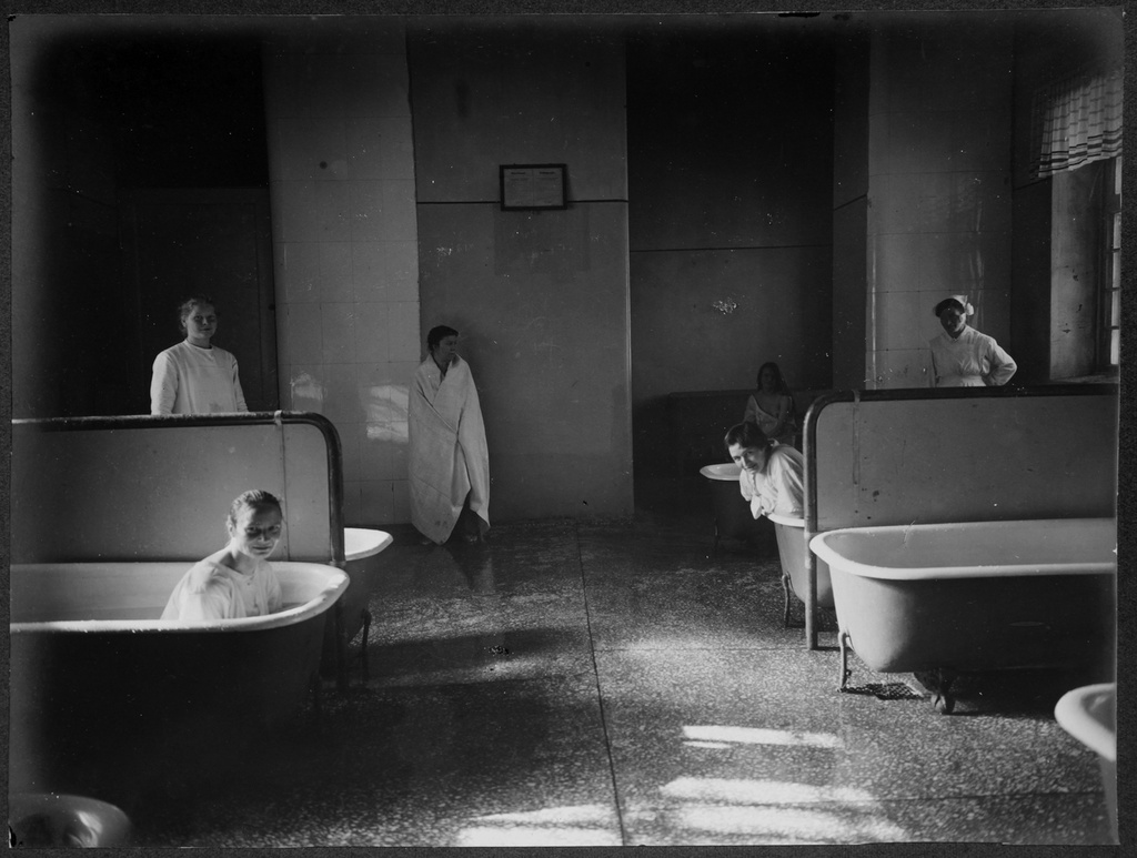 Naispotilaita kylpyhoidossa Lapinlahden sairaalan suljetulla osastolla. Huoneessa on useita kylpyammeita, joista osassa on naisia kylvyssä aluspaidoissaan. Yksi potilas seisoo lattialla pyyhkeeseen kietoutuneena. Huoneessa on myös sairaanhoitaja. N. 1904-1922.
