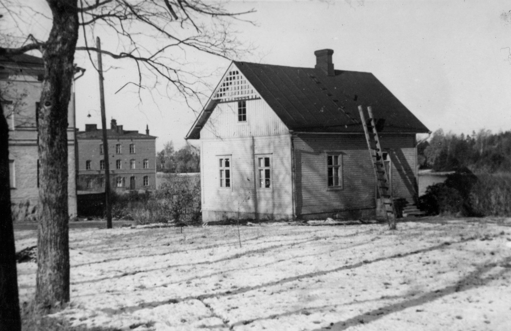 Lapinlahden sairaala-aluetta. Kuvan vasemmassa laidassa näkyy sairaalan päärakennuksen kulmaa. Taaempana rannassa häämöttää Venetsiaksi kutsuttu rakennus, jossa on toiminut mm. sairaalan pesu- ja leivintupa. Kuvan keskivaiheilla on puinen asuinrakennus, ns. Omenapuutalo. 1930-luku.
