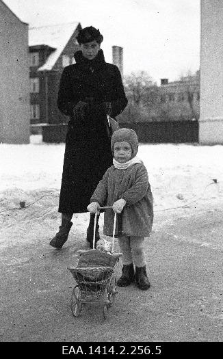 Plastic-negative woman with a small child stands in the background of buildings. The child has a small rotated dollbox.