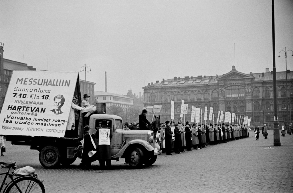 Jehovan todistaja, insinööri Kaarlo Hartevan esitelmää mainostava kulkue. Miehiä ja naisia mainos- ja tekstikylttien kanssa Rautatientorilla, vasemmalla edessä auto jonka lavalla suuri mainosjuliste.