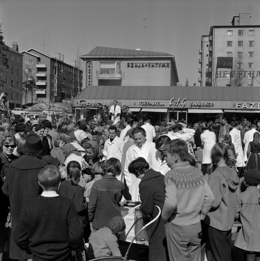 Ihmisiä Herttonimen torilla pääsiäisenä Lions clubin järjestämässä Pääsiäis munia -tapahtumassa. Taustalla arkkitehti Eliel Muoniovaaran suunnittelemat Kettutie 4:n kerrostalot sekä yksikerroksisen ostoskeskuksen ja kolmikerroksisen toimistorakennuksen muodostama Erätorin liikekeskus. Vuonna 1955 suunnitellut rakennukset valmistuivat vuosina 1956?1957. Oikealla takana elokuvateatteri Kino Herttuan mainoskyltti.