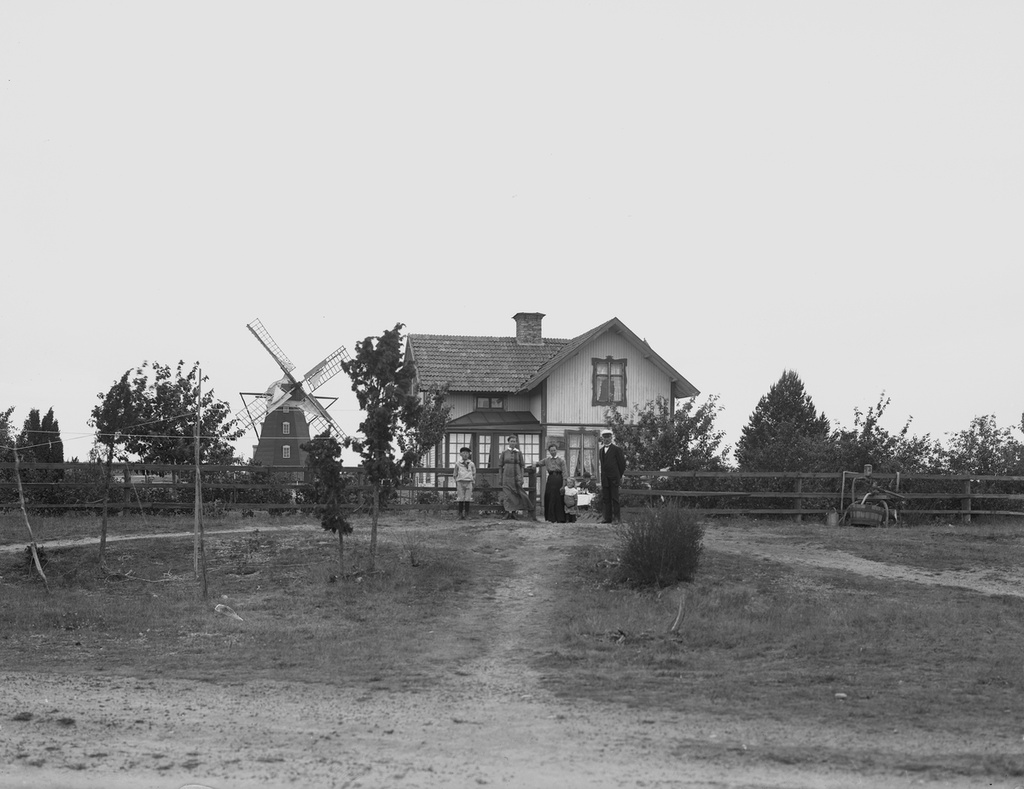 En och en halvvånings vinkelbyggt bostadshus med glasad veranda. Sex personer framför huset.
Väderkvarn i bakgrunden.
Mjölnare Pettersson -