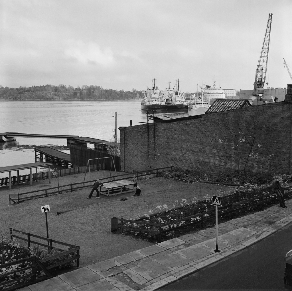 Pojat pelaavat pöytätennistä Laivastopuiston leikkipaikalla Katajanokalla. Taustalla oikealla Valmetin Katajanokan telakan nosturi ja jäänmurtajia.