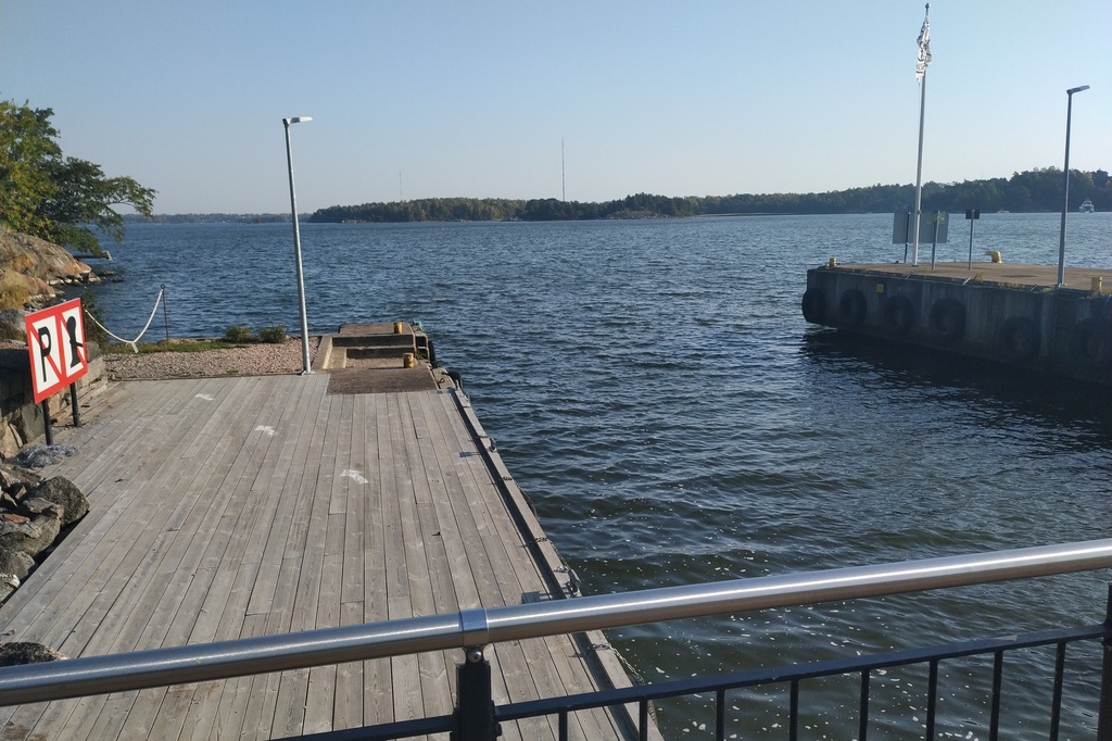 Retkeläiset menossa taksiveneeseen Vallisaaressa. Paikka on Torpedolahden laituri Vallisaaren itärannalla. Taivaanrannassa Haminansalmen toisella puolen näkyy Santahamina. rephoto