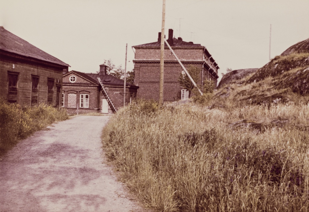 Näkymä Vallisaaresta. Oikealla on luotsitalo ja keskellä näkyy luotsitalon saunan ja pesutuvan rakennus. Vasemmalla alunperin venäläisten varuskuntansa saunaksi rakentama talo. Se on nykyään jo purettu. Tie rakennusten luo on säilynyt.