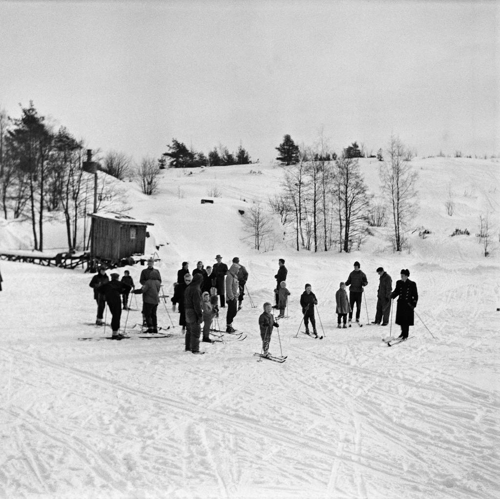 Vallisaaren lampi talvella. Jäällä on paljon ihmisiä, joista melkein kaikilla on sukset jalassa ja sauvat käsissä. Lammella järjestettiin hiistokilpailuja, joten on mahdollista, että valokuvassakin on kilpailut menossa. Ihmisjoukon takana vasemmalla näkyy pumppuhuone, joka pumppasi lammen makeaa vettä saaren asuntoja varten vesiputkiin.