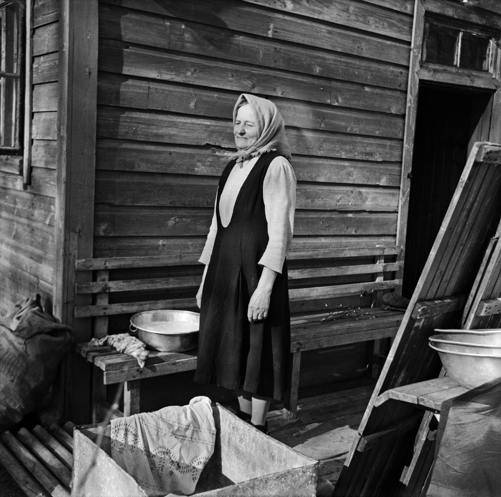 Laina Huhtala saunan edustalla Vallisaaressa. Meneillään on saunan siivoustyöt. Seinän vierustalla on penkki ja sillä pesuvati. Penkkiä vastapäätä on pöytä, jota vasten nojaavat saunan lattian puurakenteiset kävelyalustat. Aurinko paistaa.