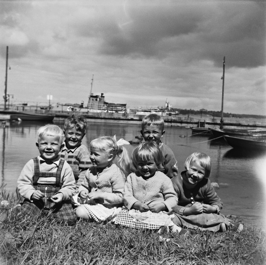 Lapsia meren rannassa nurmikolla. Lapset istuvat luotsitalon edustalla pihamaalla. Taustalla näkyy saaren satamalaituri ja yhteysalus Axel von Fersen aliturissa. Satamassa on myös puuveneitä.