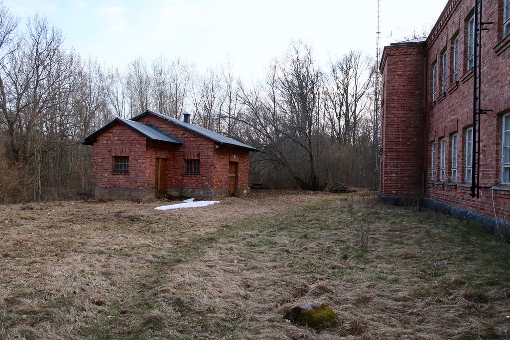 Näkymä Vallisaaren tomistotalon takapihalle. Rakennus sijaitsee Vallisaaren lammen itäpuolella. Pieni punatiilirakennus on varastorakennus, jossa on tilaa neljän perheen tavaroille. Tasainen pihamaa on paljastunut lumen alta, jota vielä on vasemmalla näkyvän pienen piharakennuksen kulmalla.