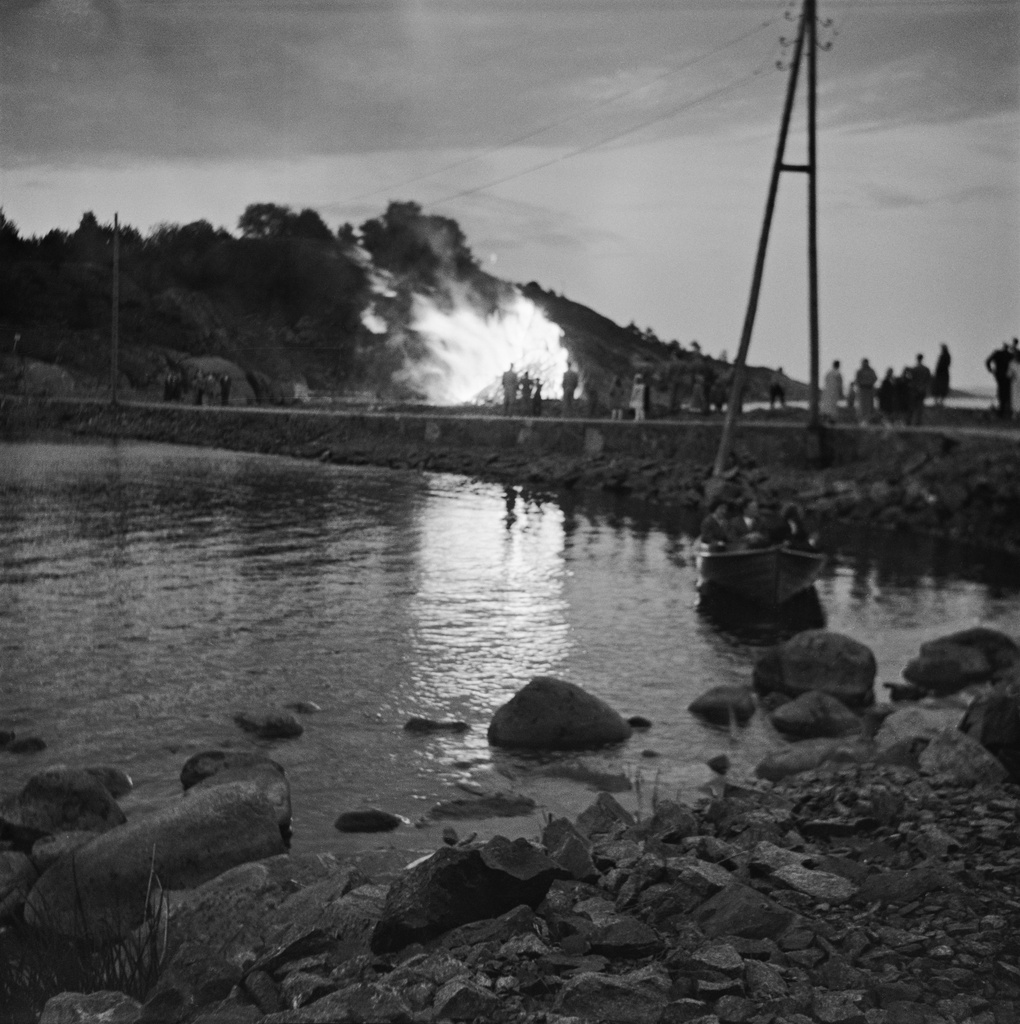 Juhannuskokko Kuninkaansaaren ja Vallisaaren välissä. Kokko on koottu saarten väliin rakennetun vallin päällä kulkevalle tielle. Valokuva on otettu Vallisaaren puolelta siten, että taustalla näkyy Kuninkaansaari. Kokon läheisyyteen on kerääntynyt ihmisiä seuraamaan roihua.