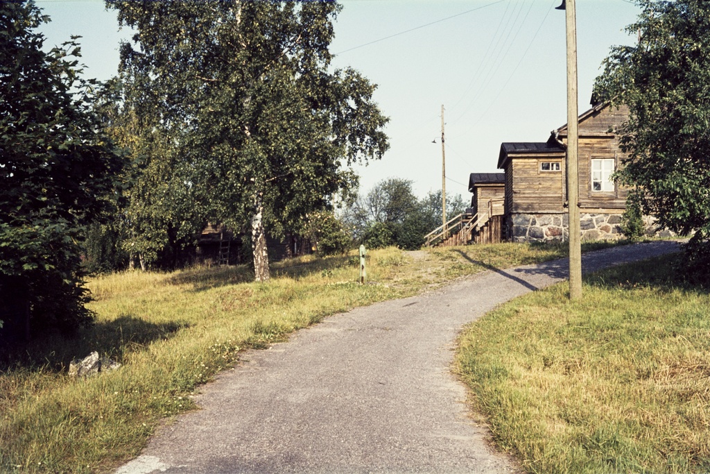 Tie ja vanha kasarmirakennus Vallisaaressa. Asfaltoitu tie johti saaren vanhalta päälaiturilta Lataamonmäelle. Kuvassa näkyvä hirsirakennus valmistui aluneprin 1860-luvlla venäläisen varuskunnan kasarmiksi. Suomen itsenäisyyden ajalla siihen tuli saaren keskussääasema. Katolle asennetut säätä havainnoivat mittarit jäävät puiden oksien taa piiloon. Rakennuksessa toimi myös koulu 1950-luvun loppupuolelle. Keskellä kuvaa tien varressa on vesiposti.