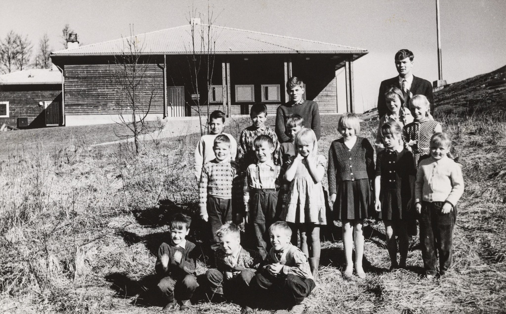 Opettaja Juha Alanen ja koululaisia mäenrinteessä Vallisaaressa. Taustalla näkyy vuonna 1958 saarelle valmistunut koulurakennus. Ryhmäkuva on otettu keväisessä auringonpaisteessa.