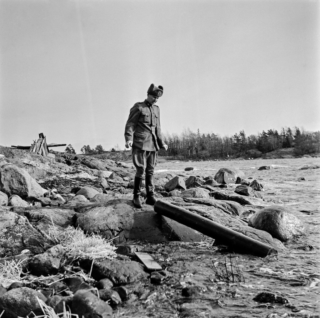 Pentti Hurskainen on löytänyt rannasta propsipuun. Se on ajautunut rantaan mereltä. Hurskainen on pukeutunut armeijan univormuun. Taustalla vasemmalla näkyvä puutavarasta kokoon kyhätty keko koostuu rantaan ajelehtineesta puusta.