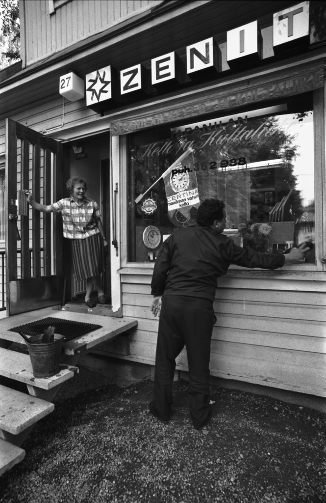 Päivöläntie 27. Mies pesemässä Tapanilan Kello ja Kultaliikkeen näyteikkunaa liikkeenhoitajan katsellessa ovella.