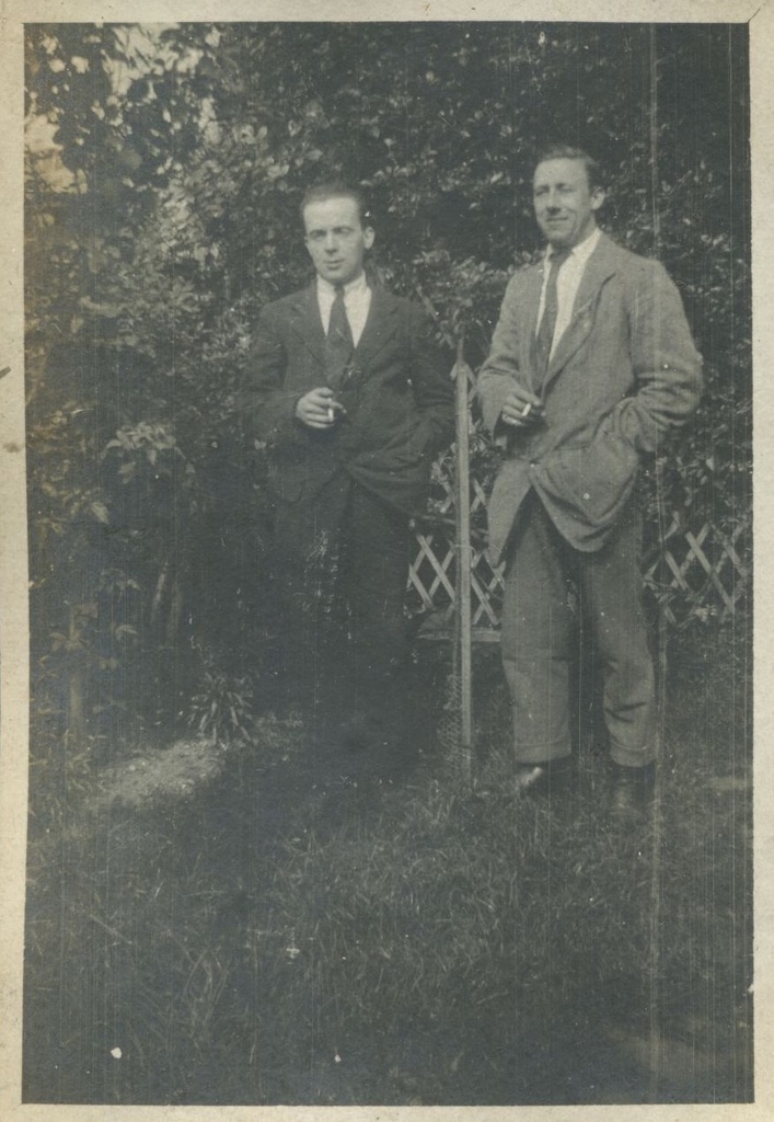 Two men in suits standing in a garden, with cigarettes in hand