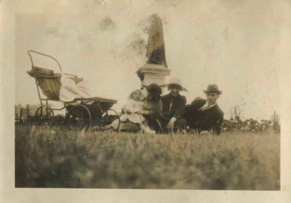 Family group seated on the ground, with a toddler and pram, in front of a statue