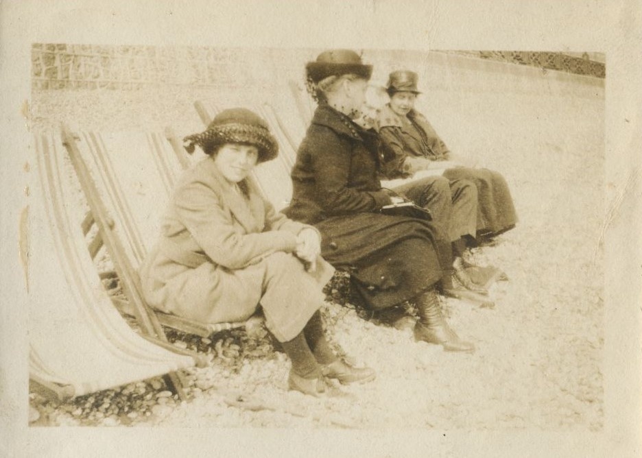 Young girl looking at the camera with two women talking behind her, all seated in deckchairs on the beach