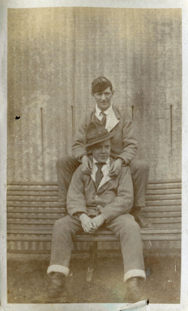 Man seated on a bench in New Zealand hat with another man seated behind him on the top rail. Tin Town, Brockenhurst.