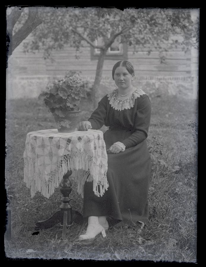 Naine aias, poseerib ümmarguse laua kõrval, millel potilill, taustal näha majasein.