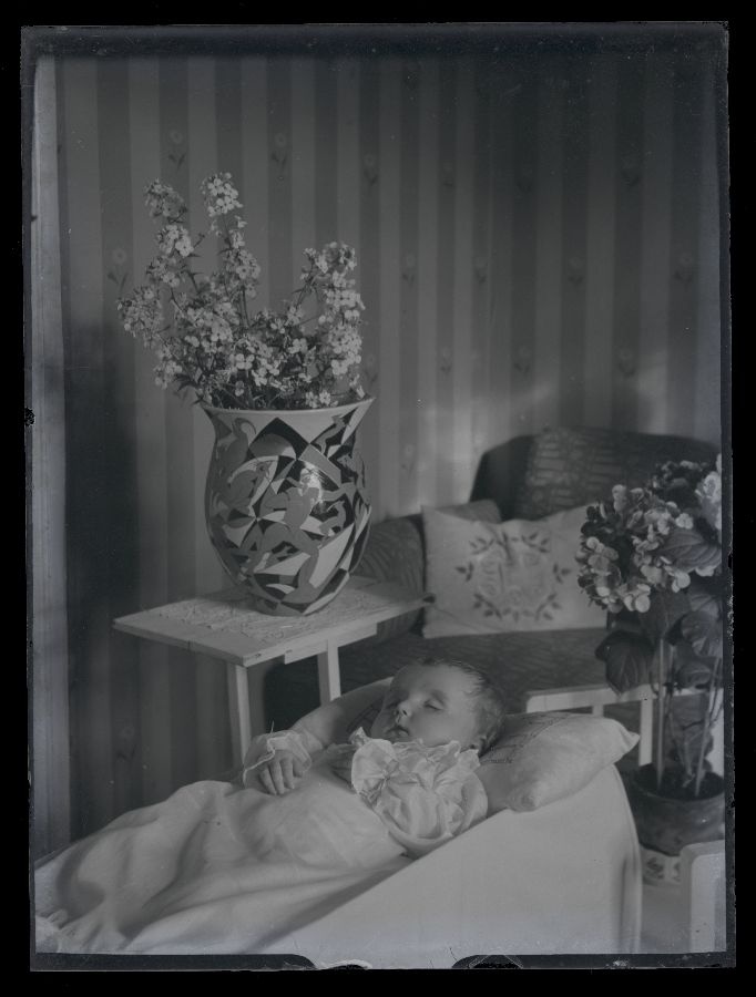 Matusefoto, väike laps avatud kirstus, kirstu taga postamendil lillevaas lilledega.