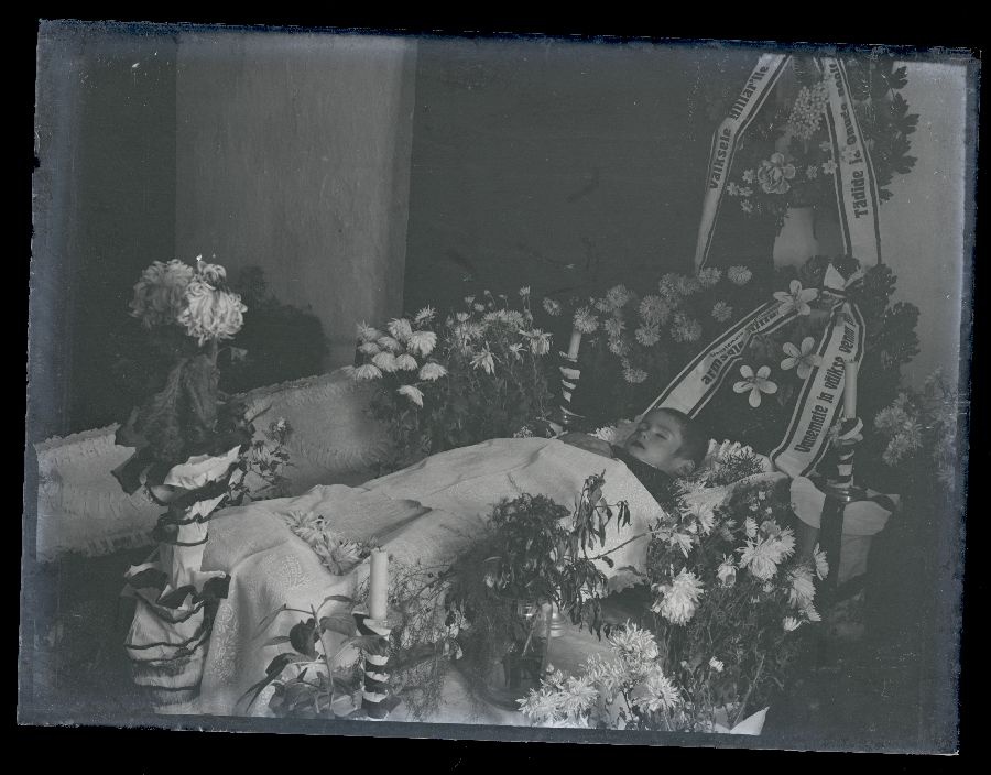 Matusefoto, väike laps avatud kirstus, kirst ümbritsetud lillede ja küünaldega.