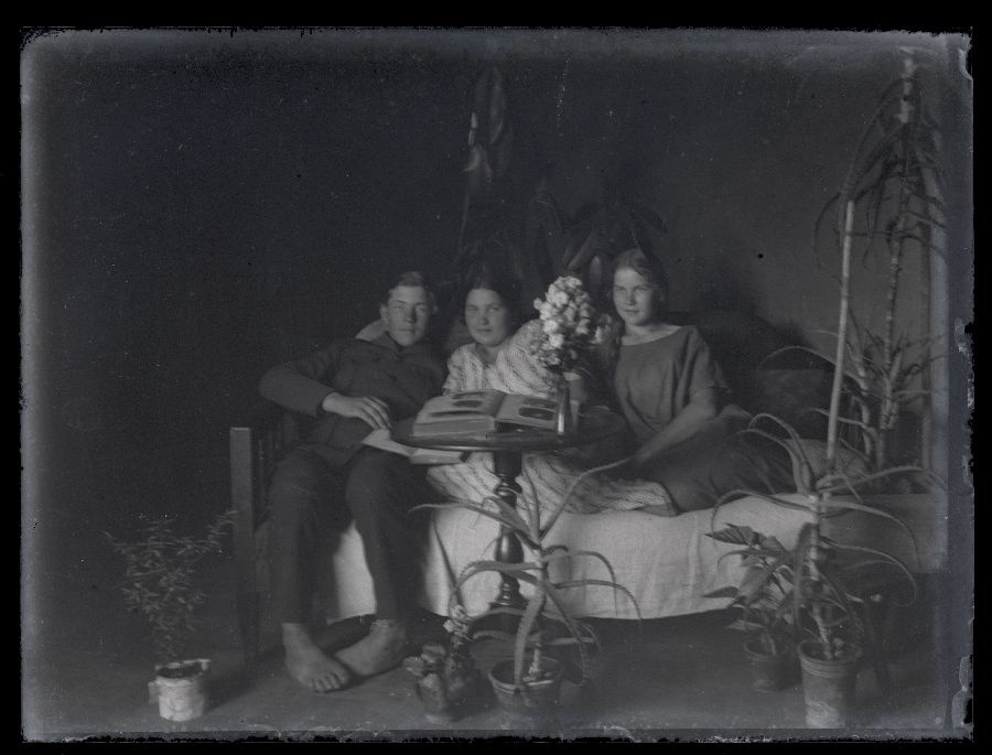 Grupifoto, kaks tüdrukut ja üks poiss istuvad diivanil, ees lauake albumi ja lillevaasiga, põrandal potililled.