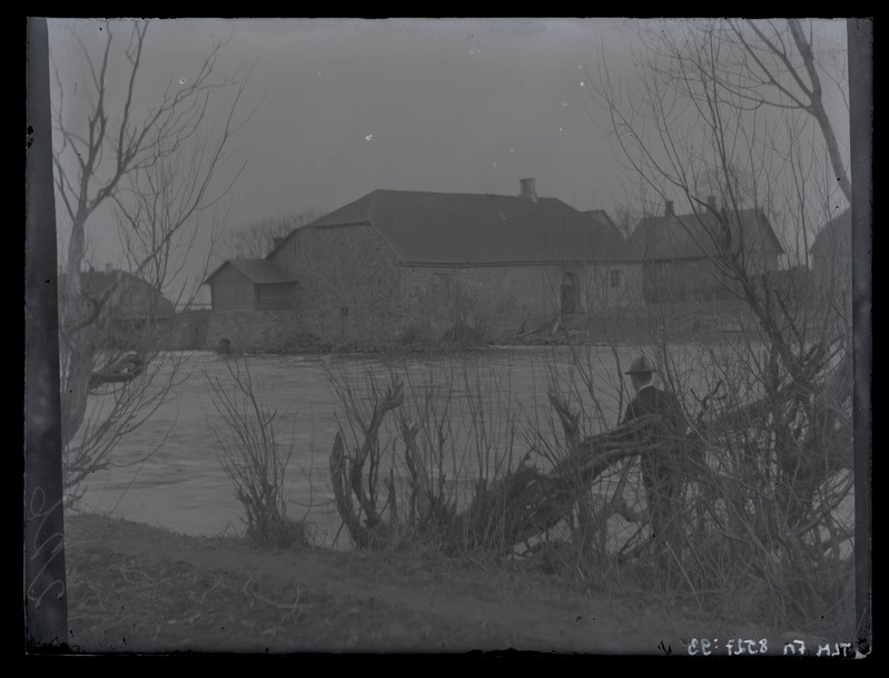Maastikuvaade vesiveskile kõrvalhoonetega, esiplaanil meesfiguur veekogu ääres, umbes 1910. aastast.