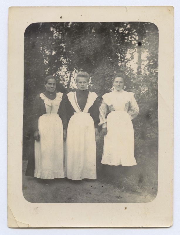 Kolm valgetes, volangidega põlledes teenijannat. Taustal puud.