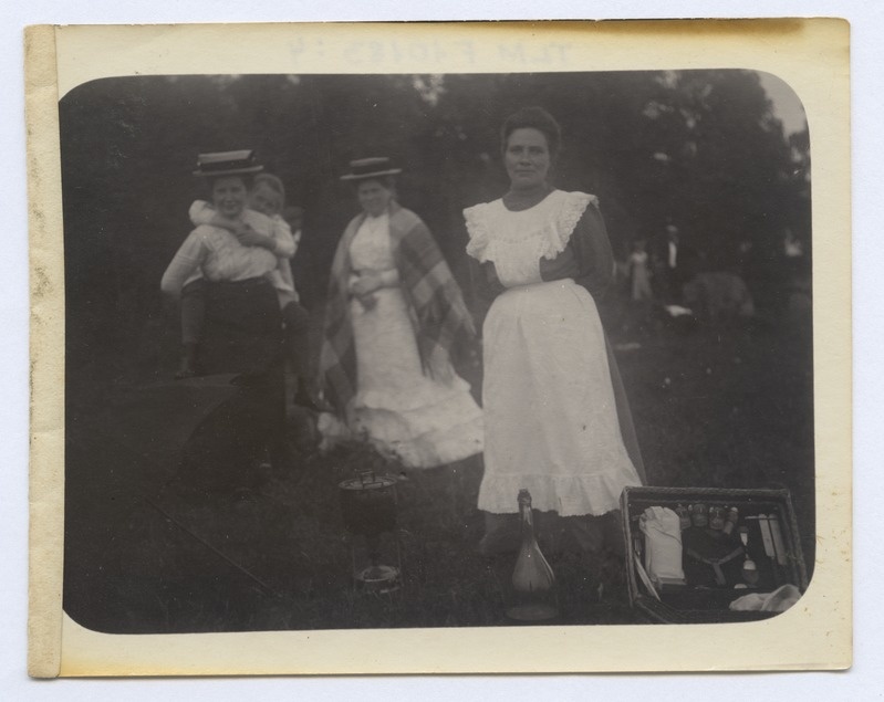 Väljasõit rohelisse. Esiplaanil valge põllega teenijatüdruk, tema ees priimus kastruliga, pudel ja kohver söögiriistadega.