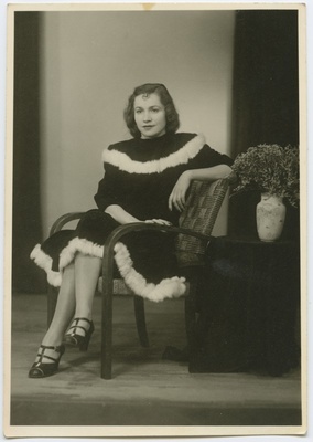 Tumedas, valge nahaga ääristatud kleidis naine leentoolis, kõrval lauake lillevaasiga. Täisportree. Ateljeefoto.  duplicate photo