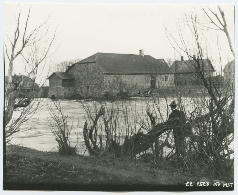 Maastikuvaade vesiveskile kõrvalhoonetega, esiplaanil meesfiguur veekogu ääres, umbes 1910. aastast.