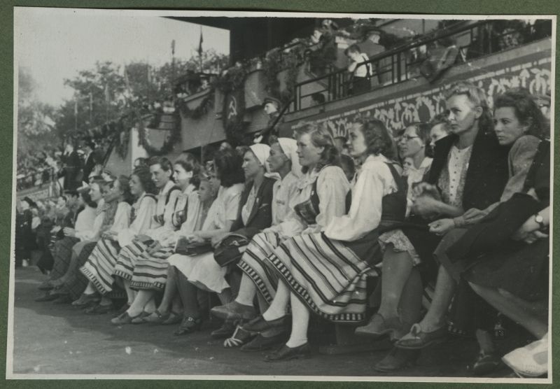 XII üldlaulupidu 28.-29. juunil 1947, Tallinnas.

Rahvariietes naised ja pealtvaatajad istumas tribüüni ääres rahvakunstiõhtul 12. üldlaulupeo eelõhtul Kadrioru staadionil.
Tribüün on kaunistatud ning sellelt ripub alla sirbi ja vasaraga lipp.