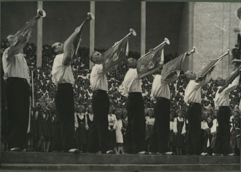 XII üldlaulupidu 28.-29. juunil 1947, Tallinnas. 
7 mustadesse viigipükstesse ja valgetesse särkidesse riietatud meest puhuvad fanfaare, et kuulutada 12. üldlaulupeo algust. 
Kõikidel fanfaaridel on samasugused vimplid, millel peal erinevad kujutised, millest üks on kannel. 
Taga paistavad laulukaare alla rivistunud koorilauljad. Paistab natukene Tallinna vana laululava seina ja mõned sambad.