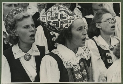 XII üldlaulupidu 28.-29. juunil 1947, Tallinnas.
Kolm rahvariietes naiskooride liiget esinemas 12. üldlaulupeol Tallinna lauluväljakul. Kõikide kolme naise rahvariideid ehivad sõled. Taustal näha teisi naiskooride lauljaid.  duplicate photo