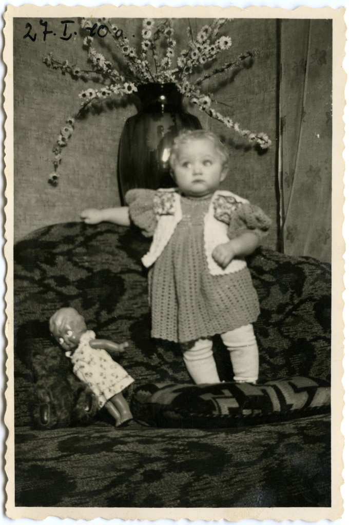 Väikelaps nukuga. Umbes 1 aastane väikelaps kootud kleidis kootud vestiga, seismas diivanil, tema jalgade juures nukk