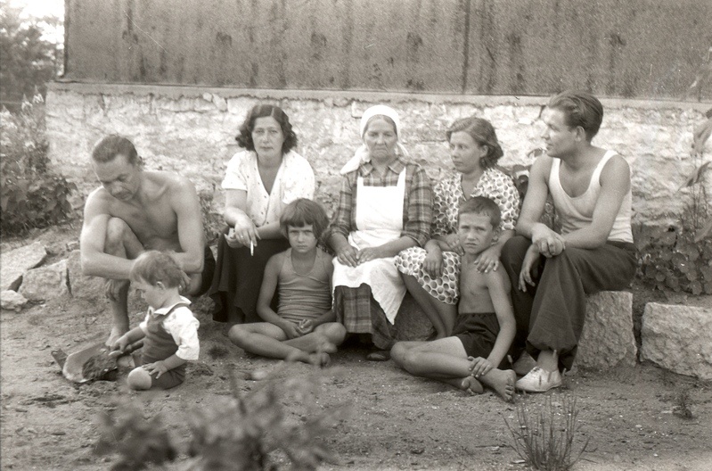 Kaks meest ja kolm naist kolme lapsega hoone taustal kividel istumas (Siina rahvas)