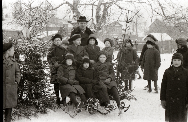Seitse poissi kahe kuuse vahel seismas ja istumas tõukekelkudel, mees nende seljataga seismas (Poeglastekooli õpilased Tartu Peetri kiriku juures asuval liuväljal)