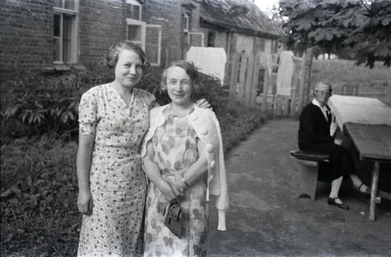 Kaks naist maja taustal seismas, taamal naine laua taga istumas