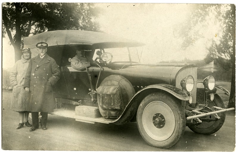 Sõiduauto, juht roolis, auto kõrval sõiduprillidega mees ja naine.