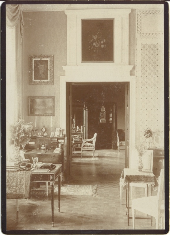 Alatskivi. Interior of the manor. In the middle of the open door; see the adjacent room. On the left side of the door, a desktop with writing bills; a landscape view and a portrait on the wall. In front of the writing table on the left table of books, vase etc. On the right small table with vase, two chairs on the edge.