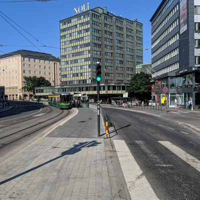 Kevättalvinen katunäkymä Hämeentieltä. Katu kääntyy vasemmalle kohdassa, jossa Vilhonvuorenkatu liittyy siihen. Risteyksen rakennus on osoitteessa Hämeentie 33 ja siitä vasemmalla Hämeentie 35. Etualalla oikealla lippakioski ja aivan oikealla näkyy osa puhelinkioskia. Äärimmäisenä vasemmalla raitiovaunu pysäkillä. Ilma on sumuisen kostea ja pilvinen, etualalla jalkakäytävälle on pinottu vanhaa lunta ja jäälohkareita. rephoto